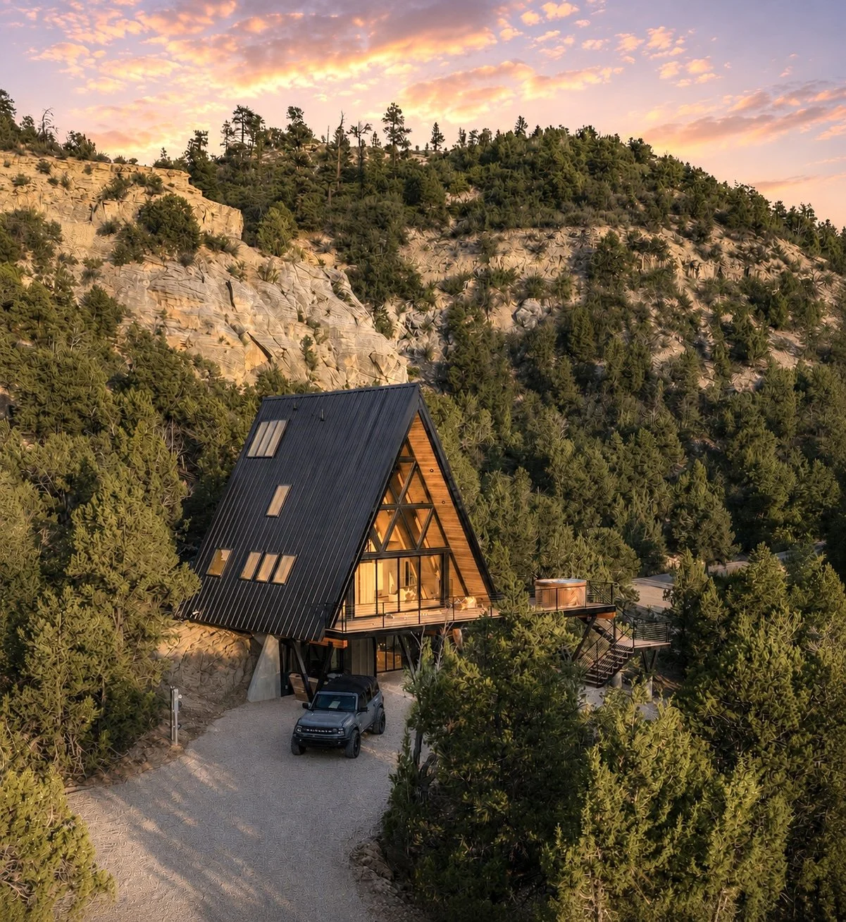 A modern A-frame house with large windows, surrounded by trees on a hillside, with a sunset sky in the background.