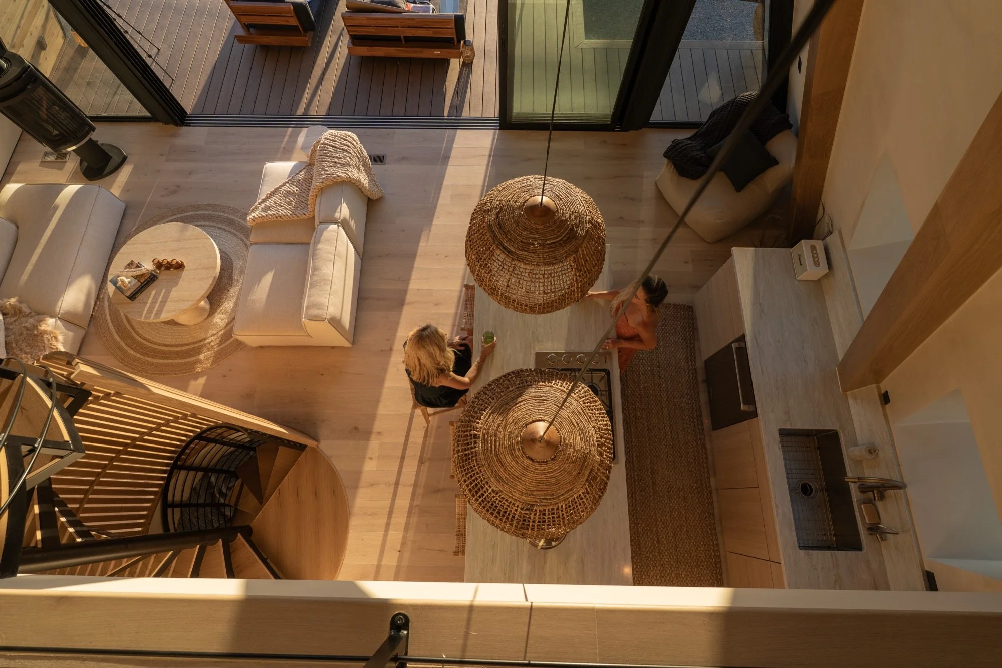 High-angle view of a modern living room with large sliding glass doors leading to an outdoor wooden deck, featuring minimalist furniture including a white sofa with beige and black cushions, a round wooden coffee table, and woven pendant lights hangi