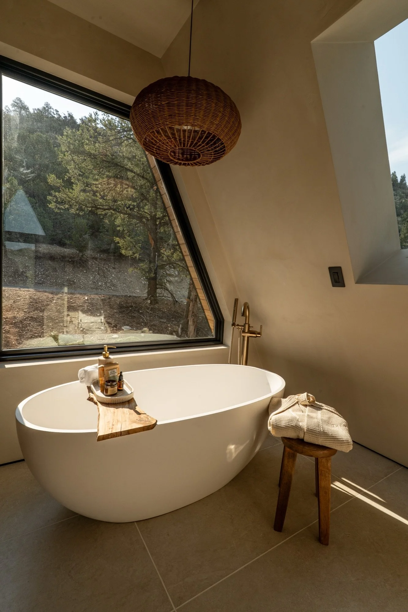 Bathroom with a white standalone bathtub positioned next to a large angled window showing a view of trees and ground outside. The tub has a wooden tray with toiletries on it. A small stool with a folded towel is beside the tub. A hanging wicker light