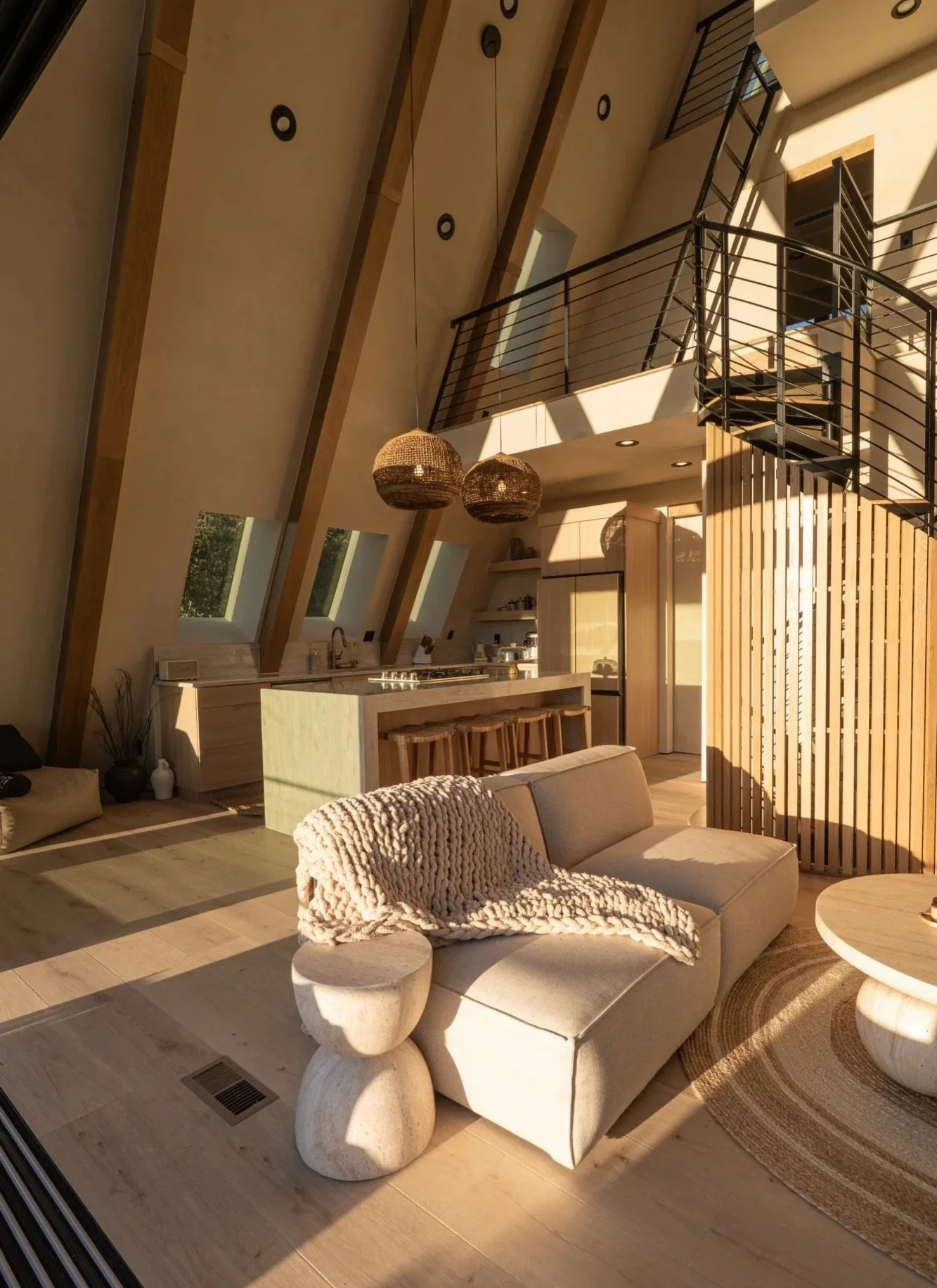 Modern living room with sloped ceilings, large windows, a beige sofa with a chunky knit throw, and a wooden kitchen island with barstools. Hanging pendant lights and a spiral staircase are also visible.