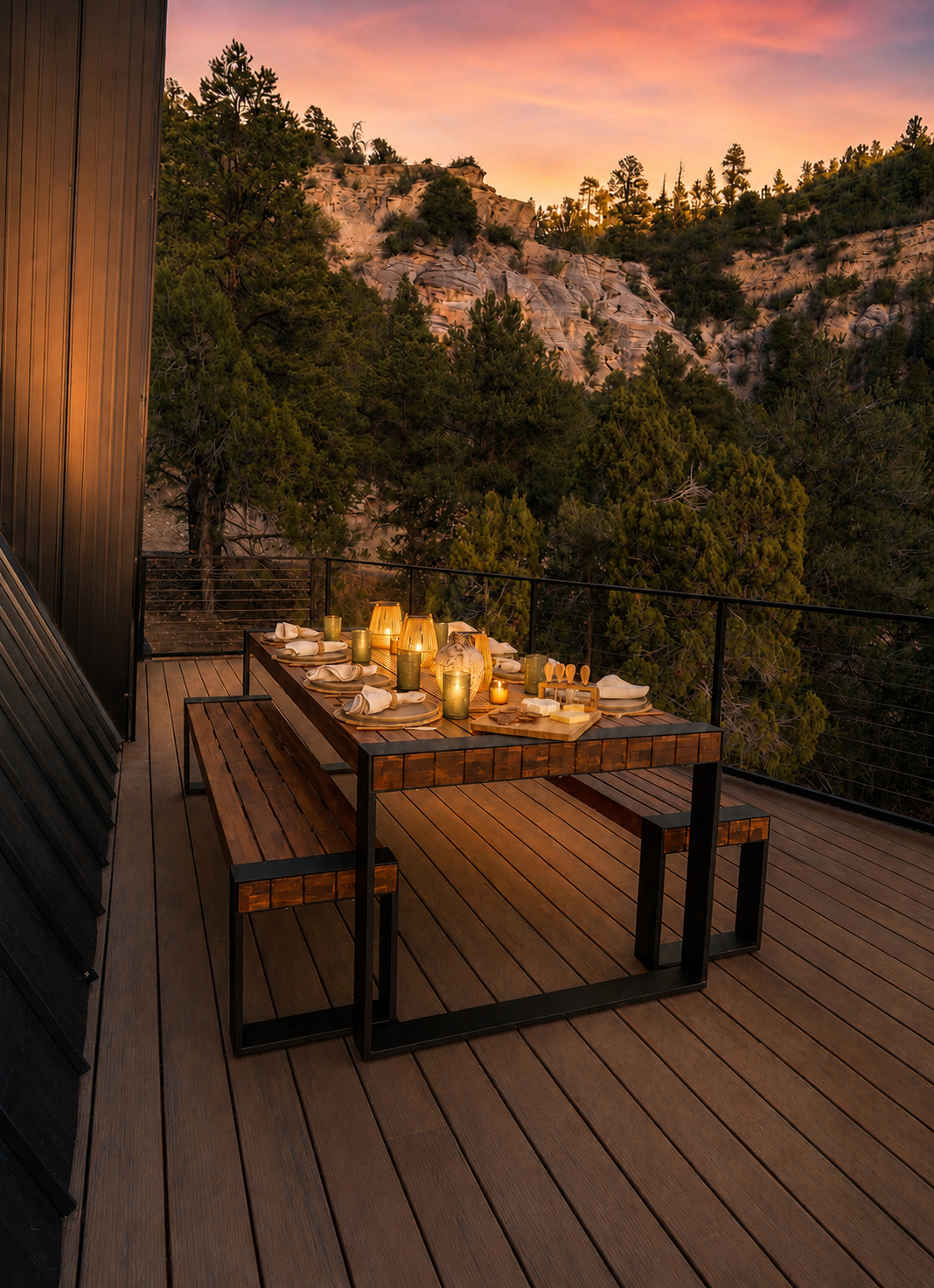Outdoor dining table set for dinner with candles and dishes on a wooden deck at sunset, overlooking a forested mountainous landscape.