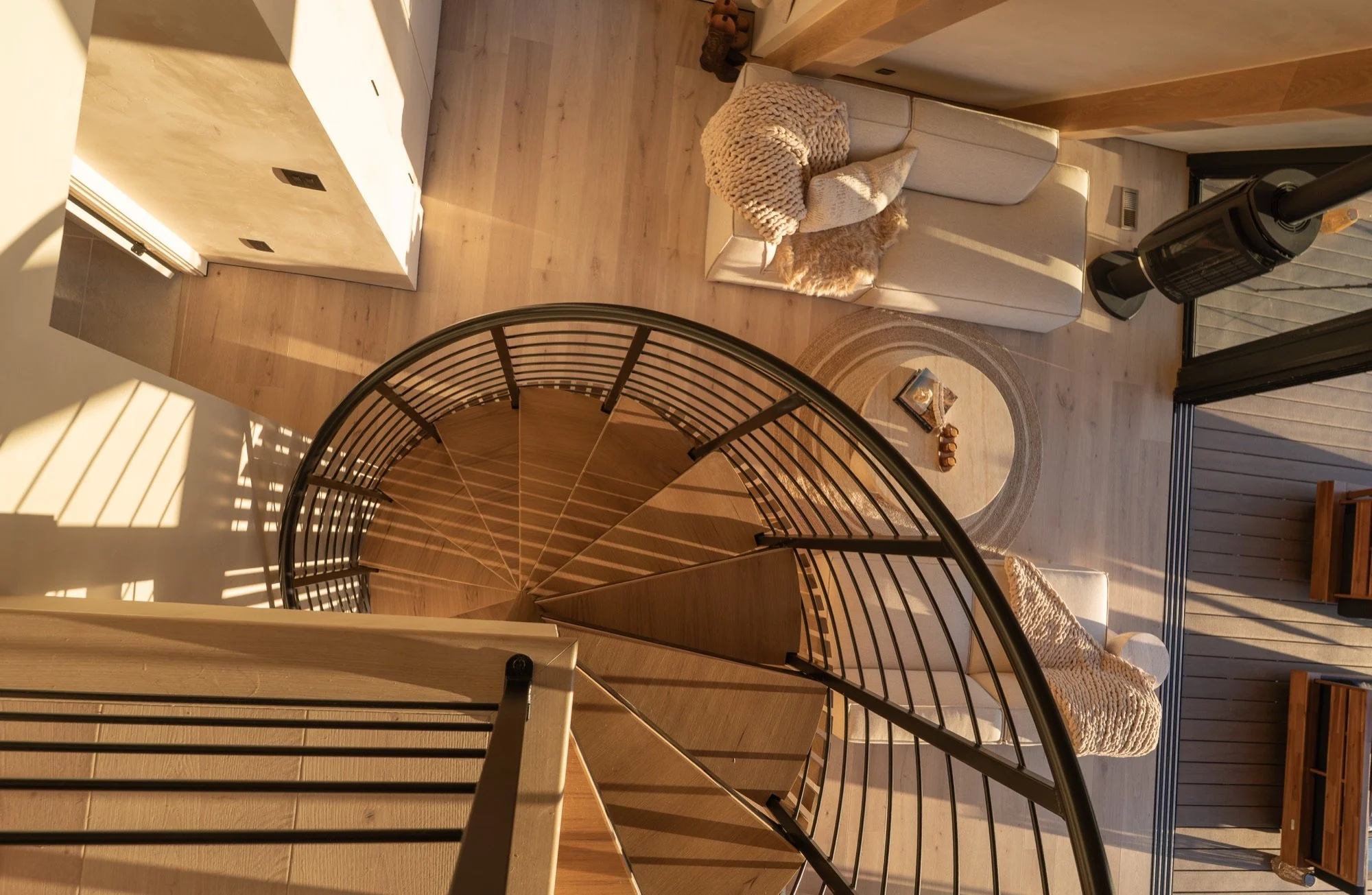 View from the top of a spiral staircase looking down into a living space with a white sofa, knitted blanket, cushions, a small circular rug, and a heater. There is a sliding glass door leading to an outdoor deck.