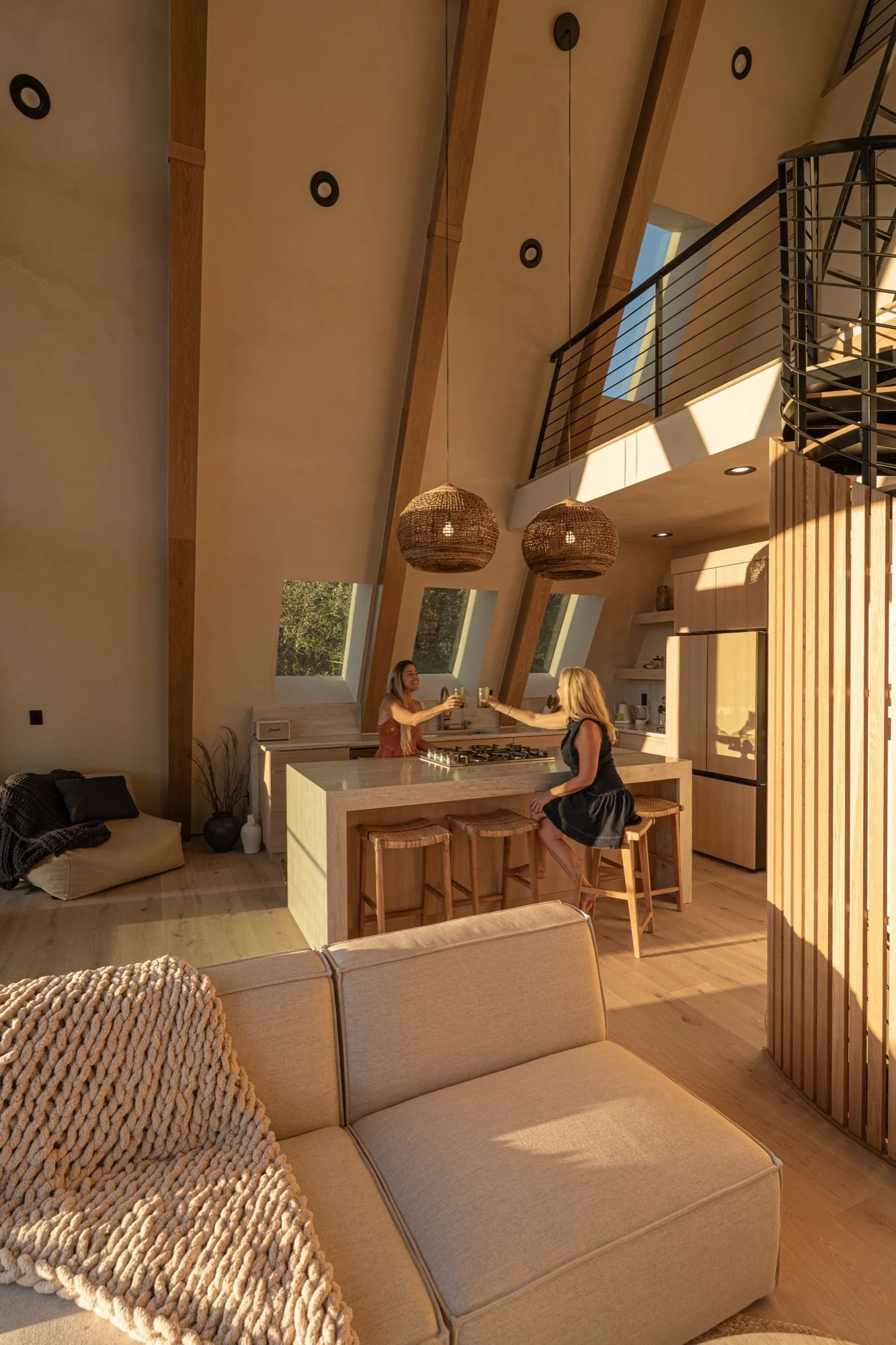 Interior of a modern, open-concept kitchen and living space with high, angled ceilings, large windows, hanging wicker light fixtures, and two women toasting with drinks at a kitchen island.