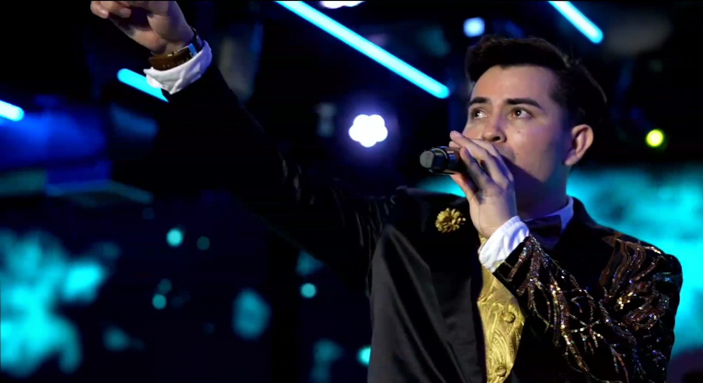 Male singer with dark hair, holding a microphone, wearing a black and gold embroidered jacket, performing on stage with blue and purple lights. La voz Guacalito, Alberto Jimenez cantante latino