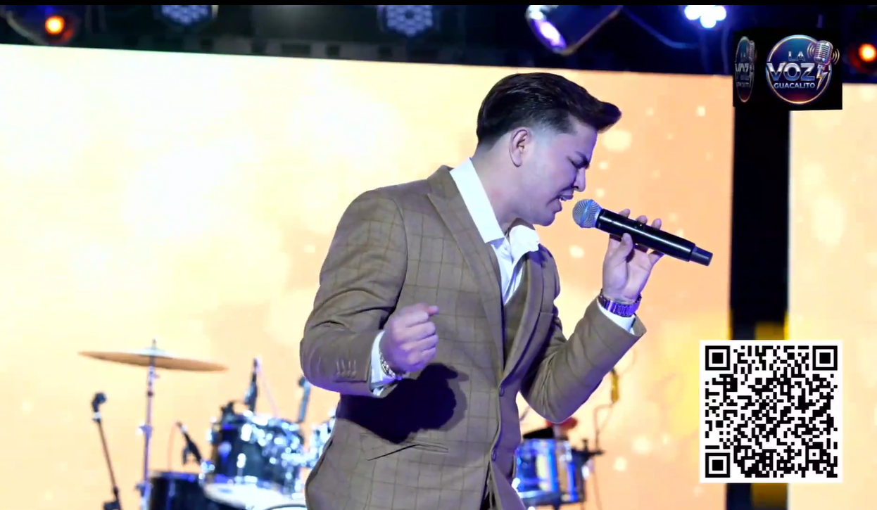 A man in a brown plaid suit is singing into a microphone on stage with a yellow background and drums in the background.El Guacalito, La Voz el Guacalito