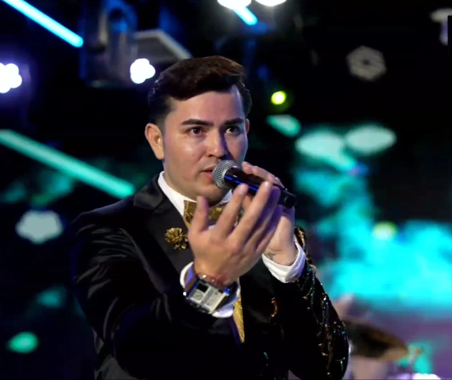 A man in a black tuxedo with gold accents is holding a microphone and singing or speaking on stage with colorful lights in the background. La voz Guacalito, Alberto Jimenez cantante latino