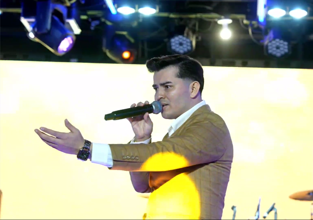 A male singer with dark hair styled back, wearing a brown suit, white shirt, and watch, performing on stage with a microphone in his right hand and gesturing with his left hand against a yellow-lit background with stage lights. El Guacalito, La Voz e