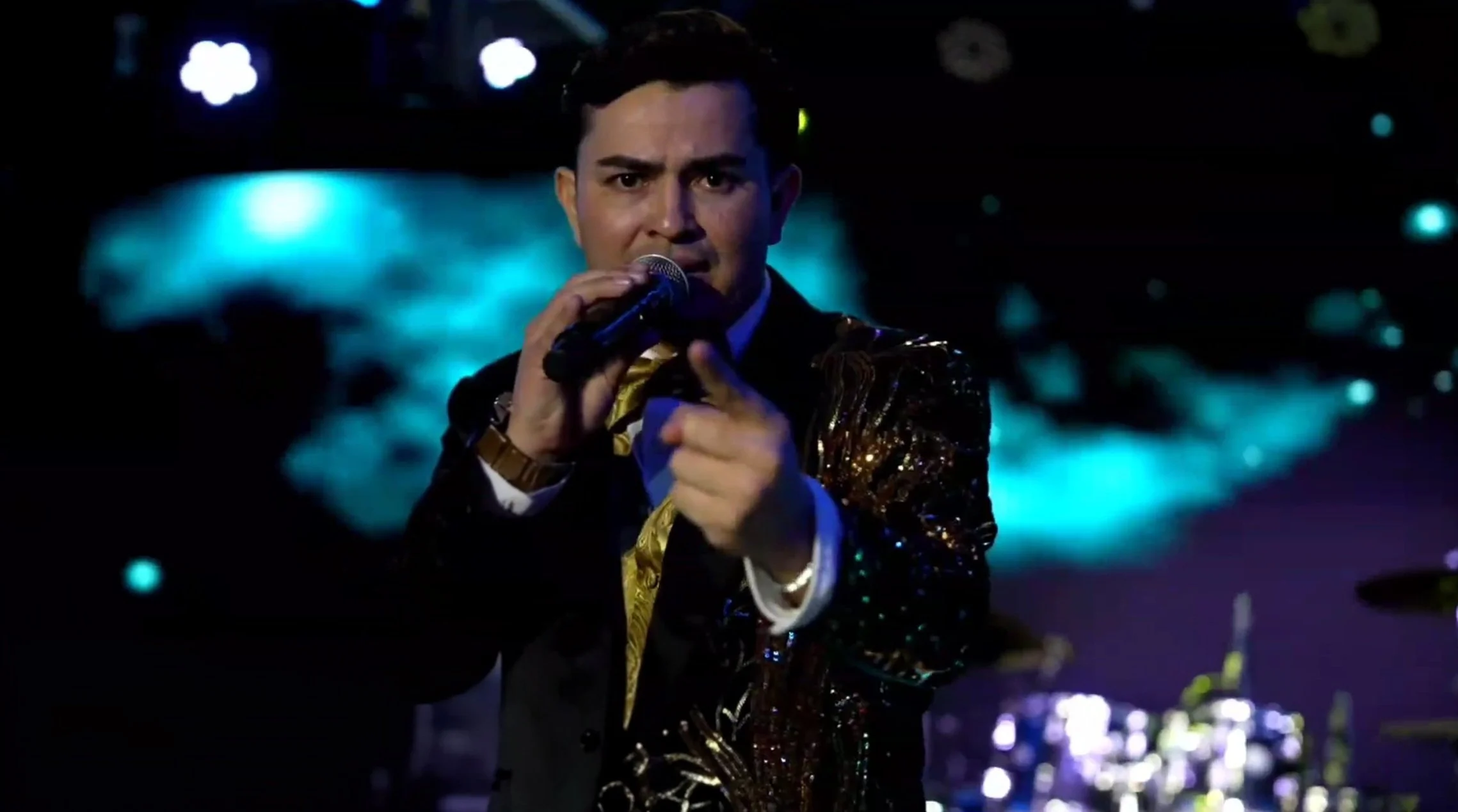 Man dressed in a shiny black and gold jacket singing into a microphone on stage with a colorful abstract background. La voz Guacalito, Alberto Jimenez cantante latino