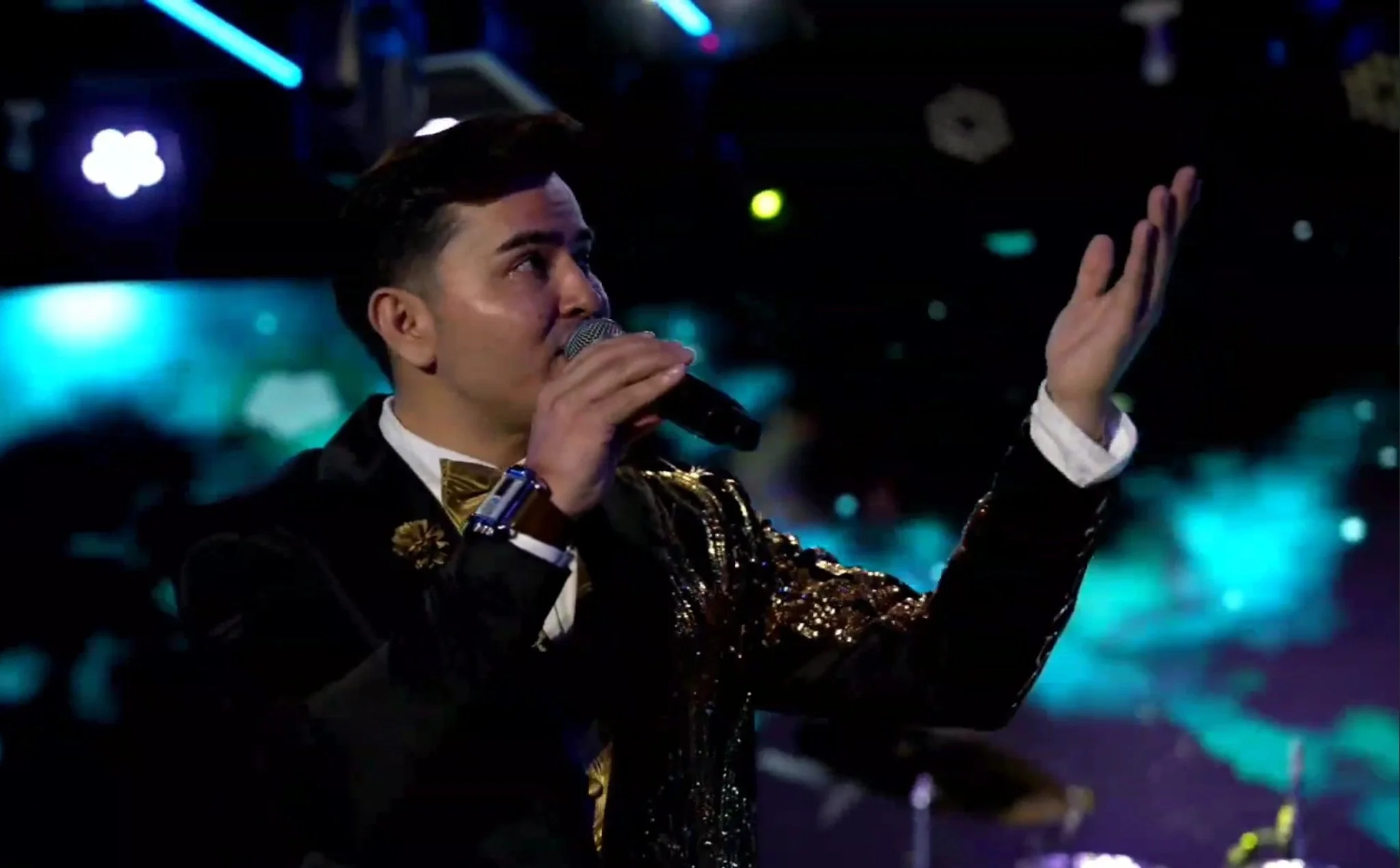 Man wearing a black blazer with gold embellishments, holding a microphone, singing or speaking on stage with colorful lights in the background. La voz Guacalito, Alberto Jimenez cantante latino