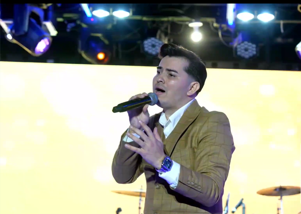 A man in a brown blazer and white shirt singing into a microphone on stage with stage lights above and a yellow screen behind him.El Guacalito, La Voz el Guacalito