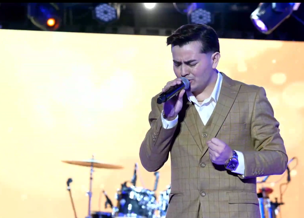 A man in a brown checkered suit singing into a microphone on stage with a yellow screen background and drums.El Guacalito, La Voz el Guacalito