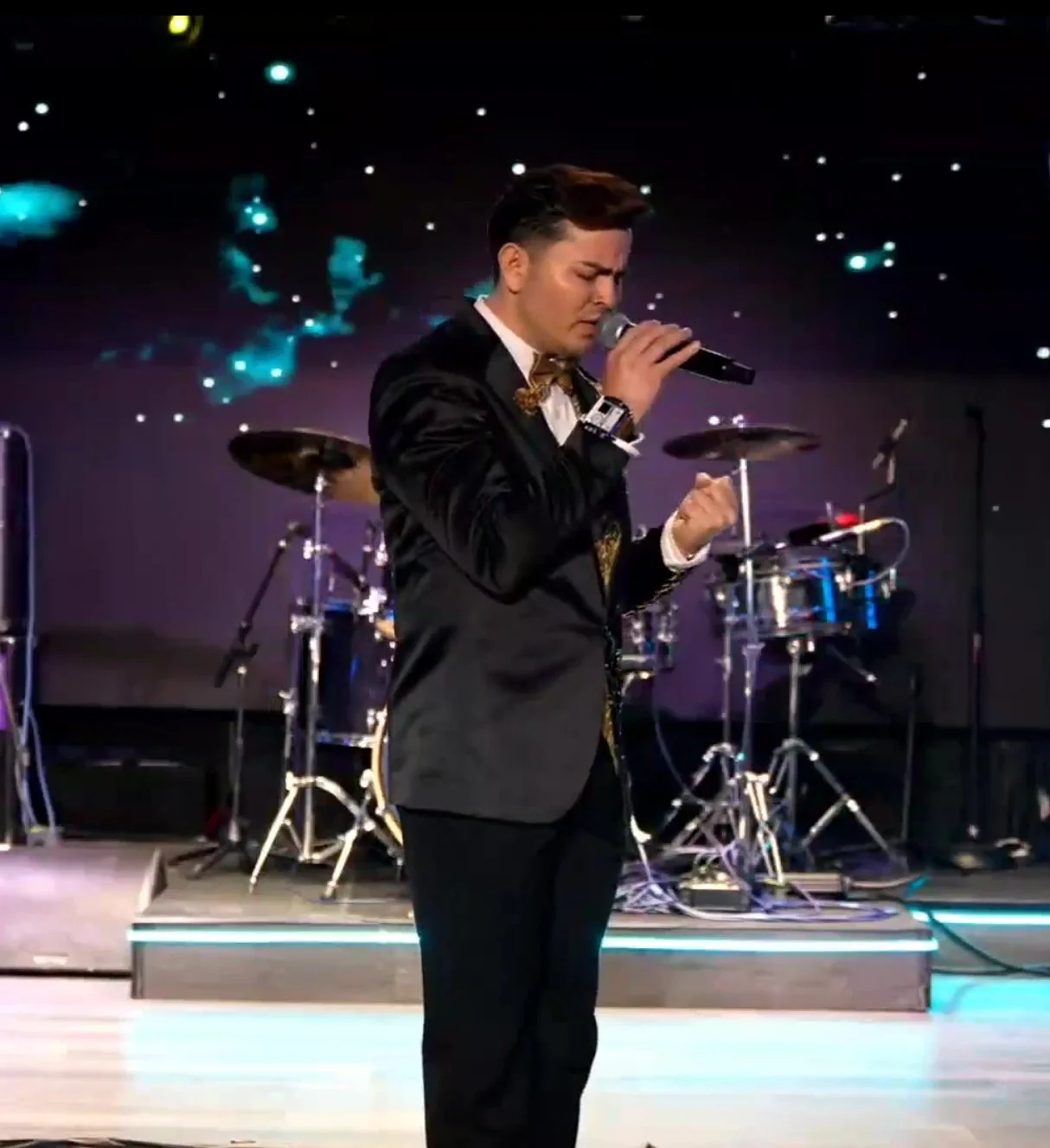 A male singer in a black tuxedo singing into a microphone on a stage with a drum set and a starry background. La voz Guacalito, Alberto Jimenez cantante latino
