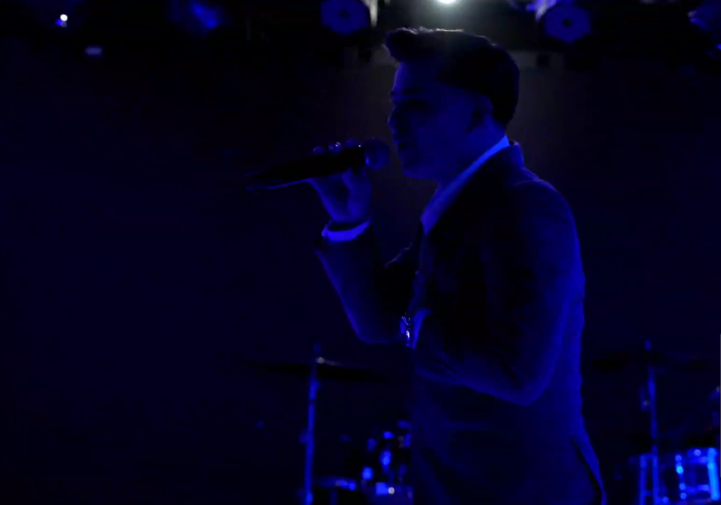 A silhouetted male singer in a suit performing with a microphone on stage, illuminated by blue stage lighting. El Guacalito, La Voz el Guacalito