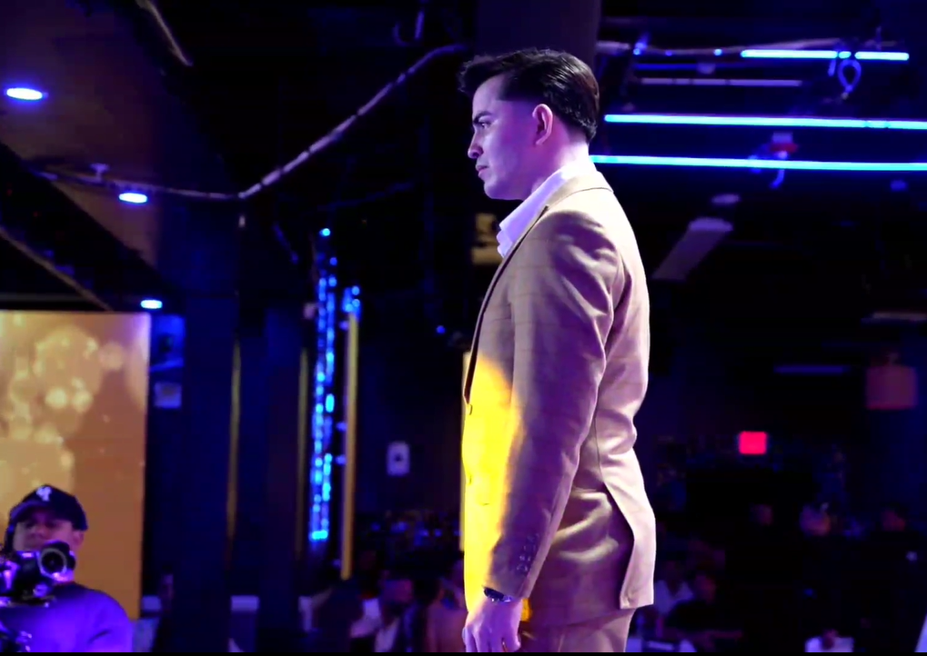 A man dressed in a beige suit standing on a stage with a serious expression, illuminated by stage lights, in front of an audience in a dark venue with blue lighting.El Guacalito, La Voz el Guacalito