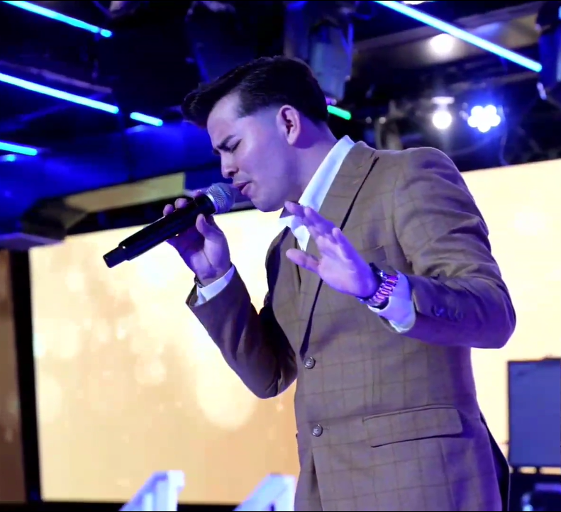 A man in a tan suit singing into a microphone with eyes closed and one hand raised, with colorful stage lights and a yellow backdrop behind him.El Guacalito, La Voz el Guacalito