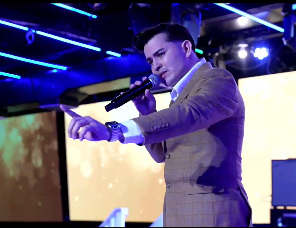 A young man in a brown checked blazer and white shirt is holding a microphone and speaking or singing on stage. The stage is lit with blue lights and has large screens in the background. El Guacalito, La Voz el Guacalito