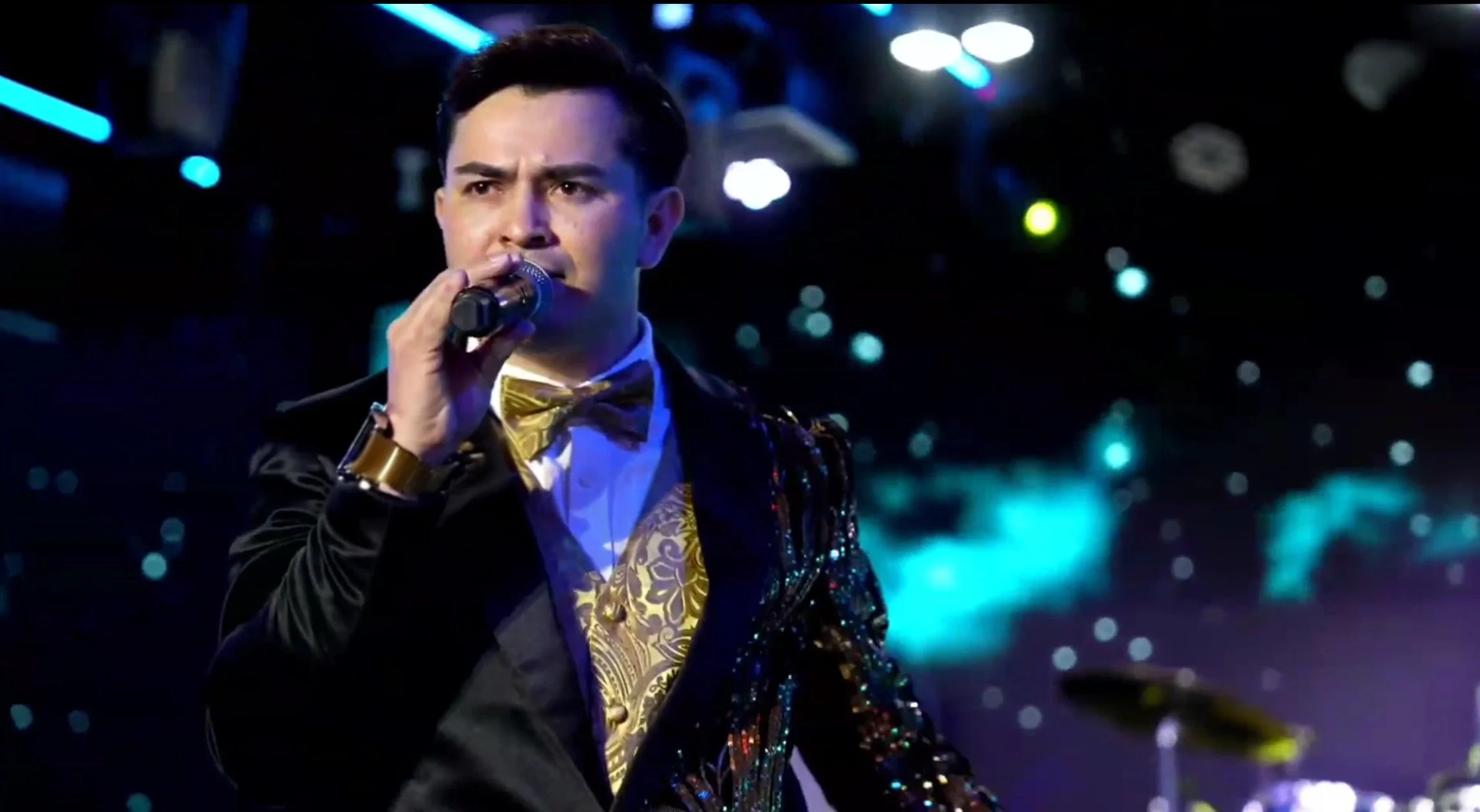 A man dressed in a shiny, ornate tuxedo with a gold bow tie and gold accents, holding a microphone and performing on stage with colorful lights and a starry background. La voz Guacalito, Alberto Jimenez cantante latino