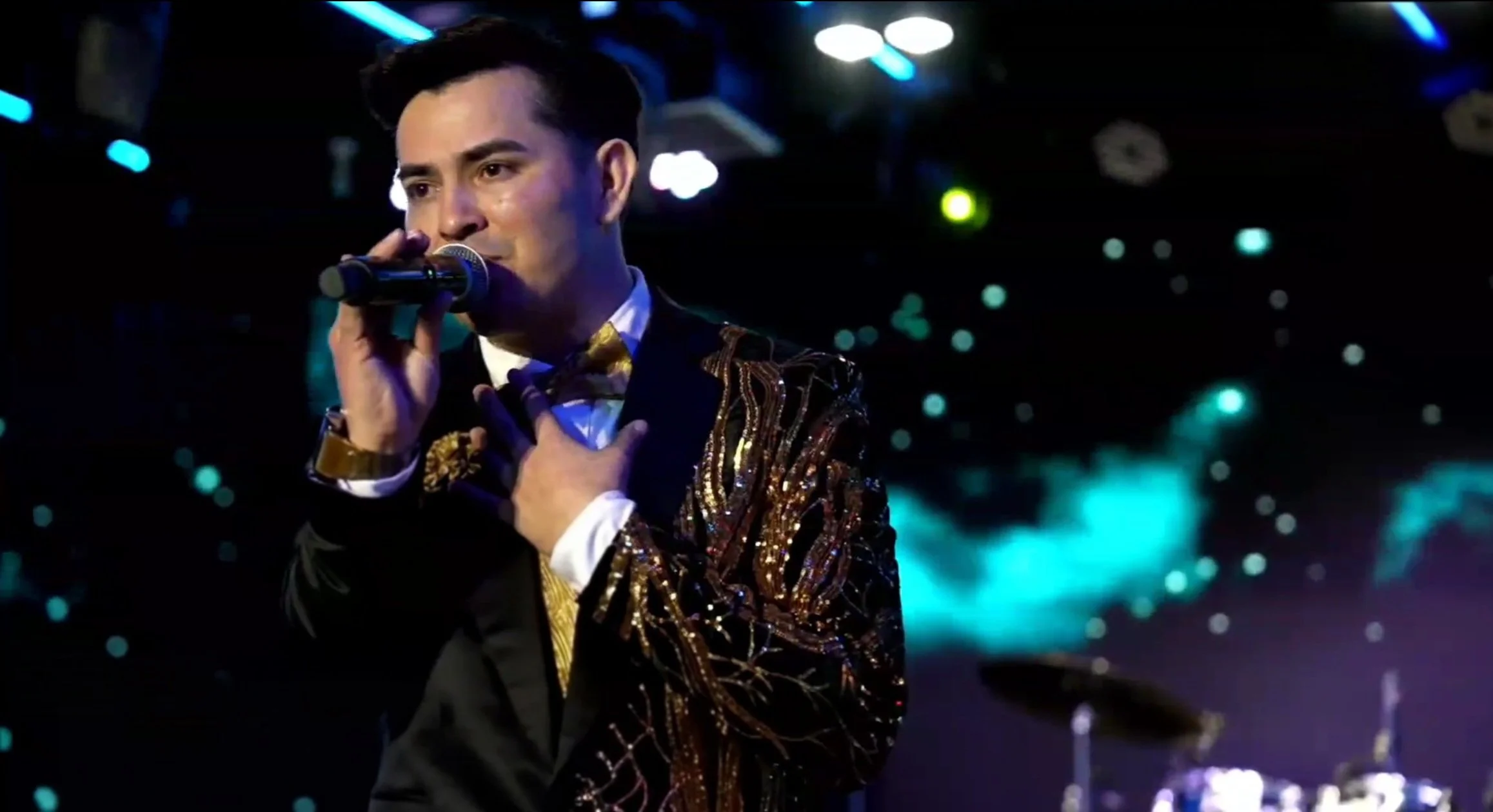 A man performing on stage with a microphone, wearing a black suit with gold and black patterned fabric, black bow tie, and white shirt, with a dark background and colorful lights. La voz Guacalito, Alberto Jimenez cantante latino