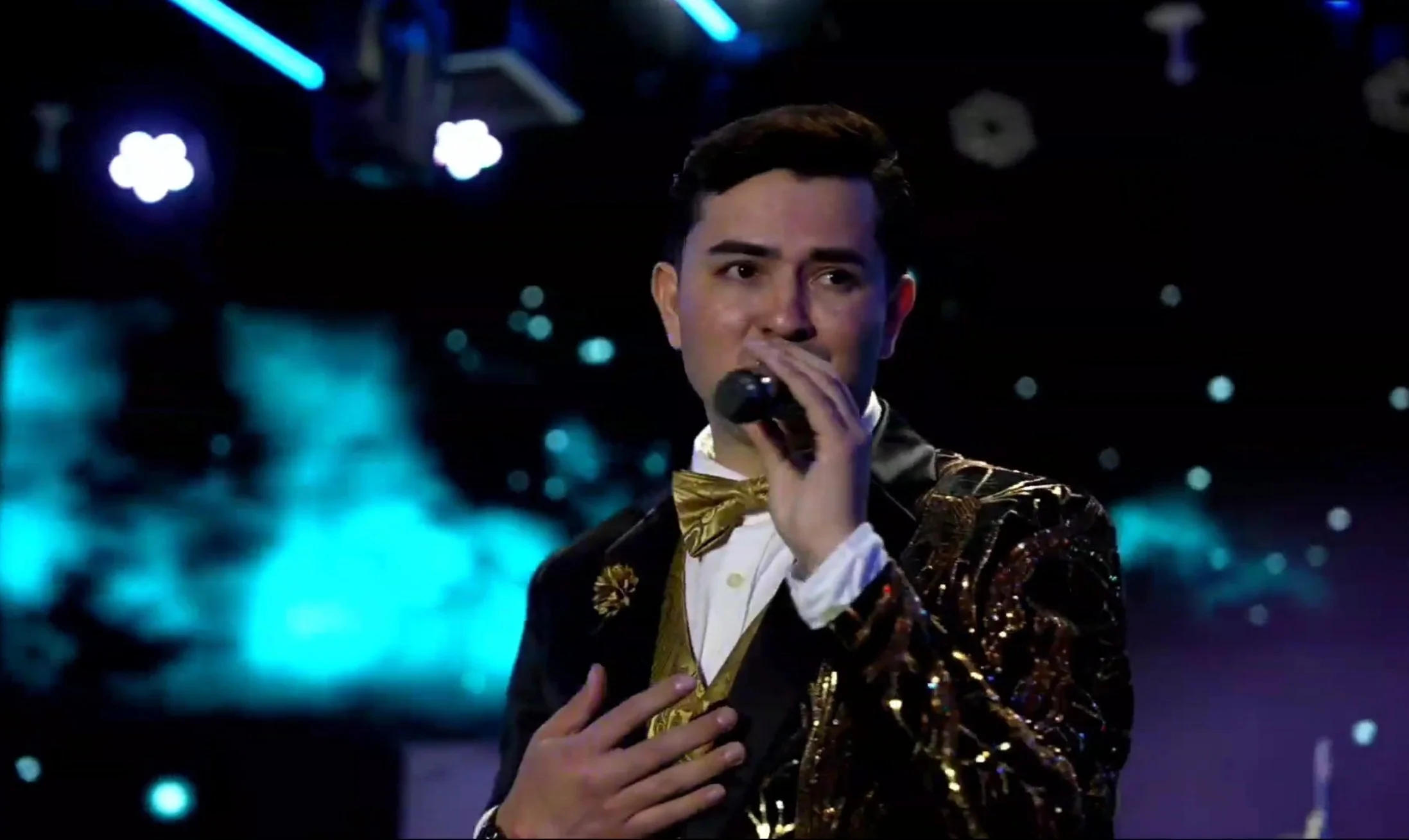A man in a shiny black and gold jacket, white shirt, and gold bow tie holding a microphone and singing on stage with a background of blue and purple lights. La voz Guacalito, Alberto Jimenez cantante latino