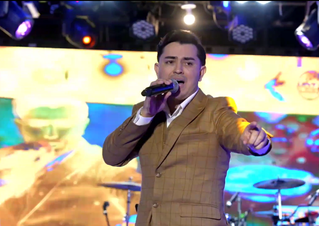 A man in a brown suit holding a microphone, performing on stage with colorful lighting and a vibrant digital background El Guacalito, La Voz el Guacalito
