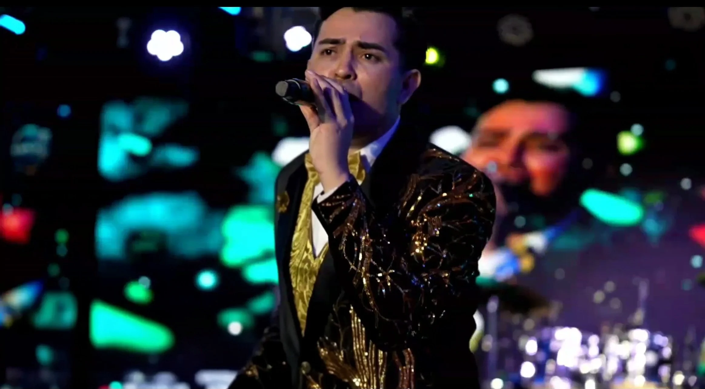A male performer singing into a microphone on stage with colorful lights and a blurred background. La voz Guacalito, Alberto Jimenez cantante latino