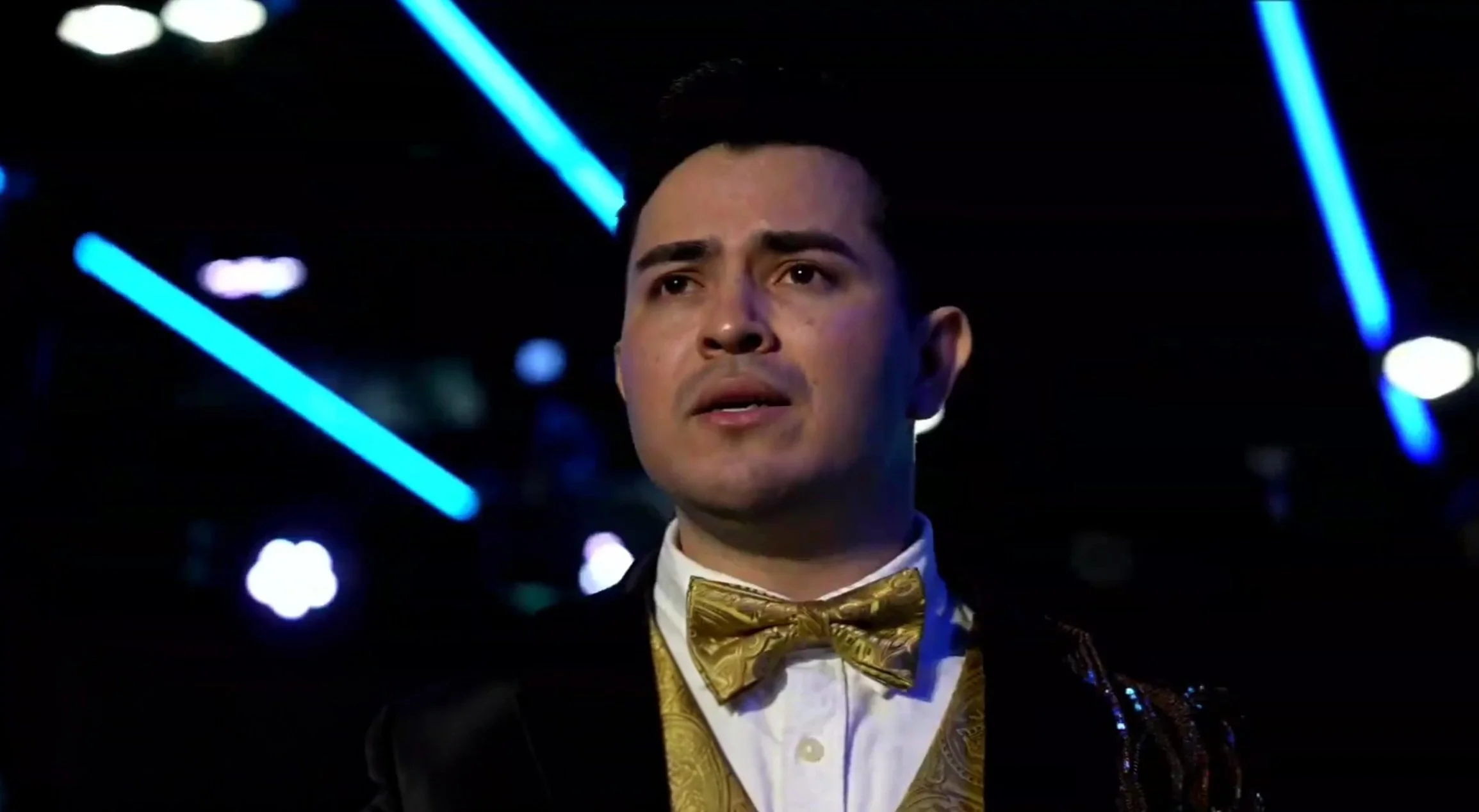 A man in formal attire, wearing a gold bow tie and a jacket with gold embellishments, stands in a dimly lit space with blue neon lights and blurred lights in the background. La voz Guacalito, Alberto Jimenez cantante latino