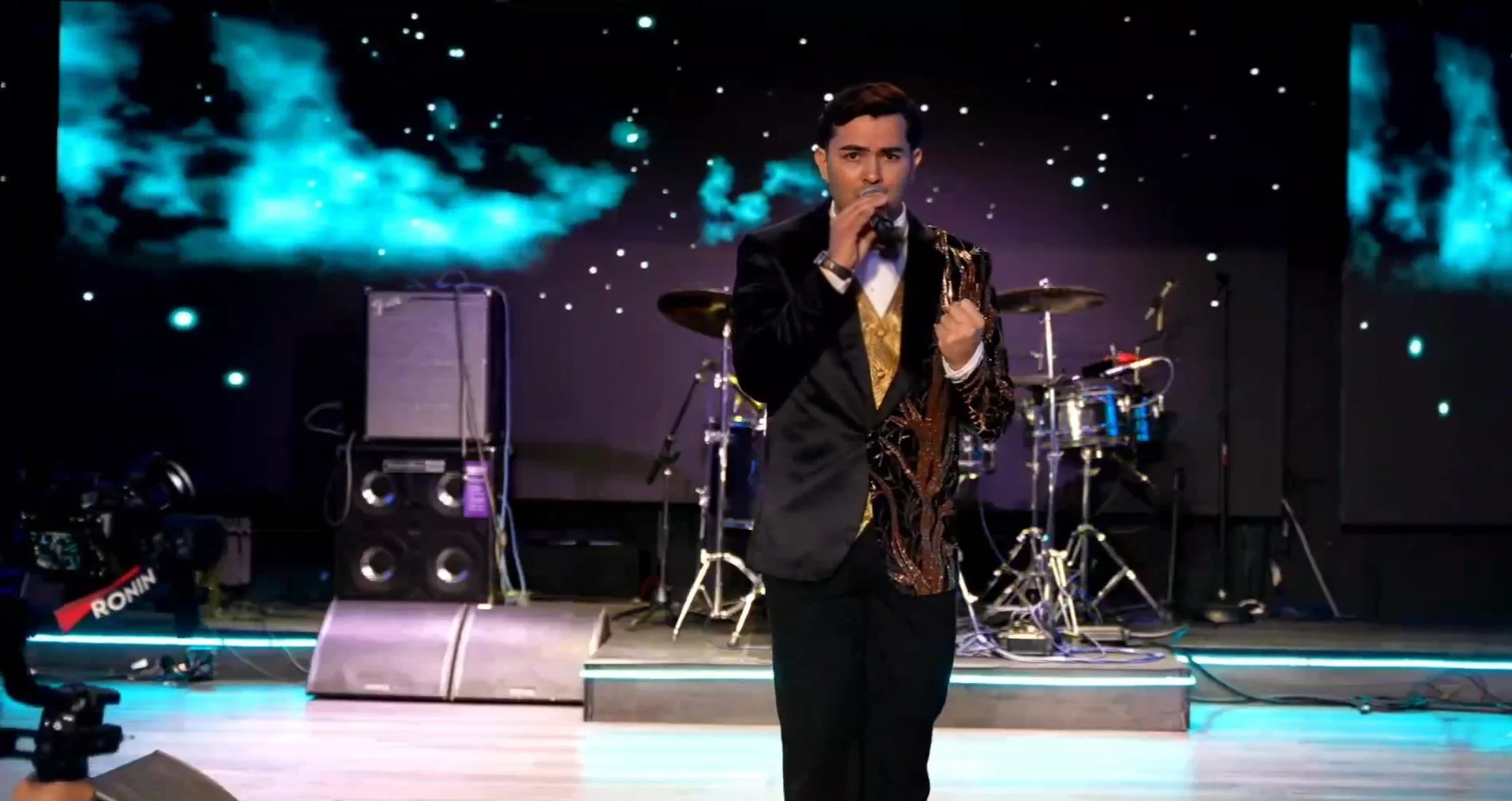 Man in tuxedo singing into microphone on stage with drum set and large screen displaying blue clouds in the background. La voz Guacalito, Alberto Jimenez cantante latino