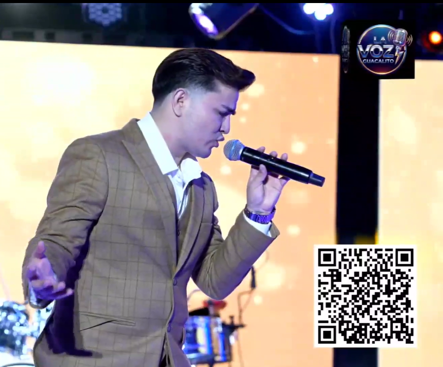 Man in a brown checked suit singing into a microphone on stage with a large glowing yellow screen behind him and a logo 'La Voz Guácalit0' in the top right corner. El Guacalito, La Voz el Guacalito