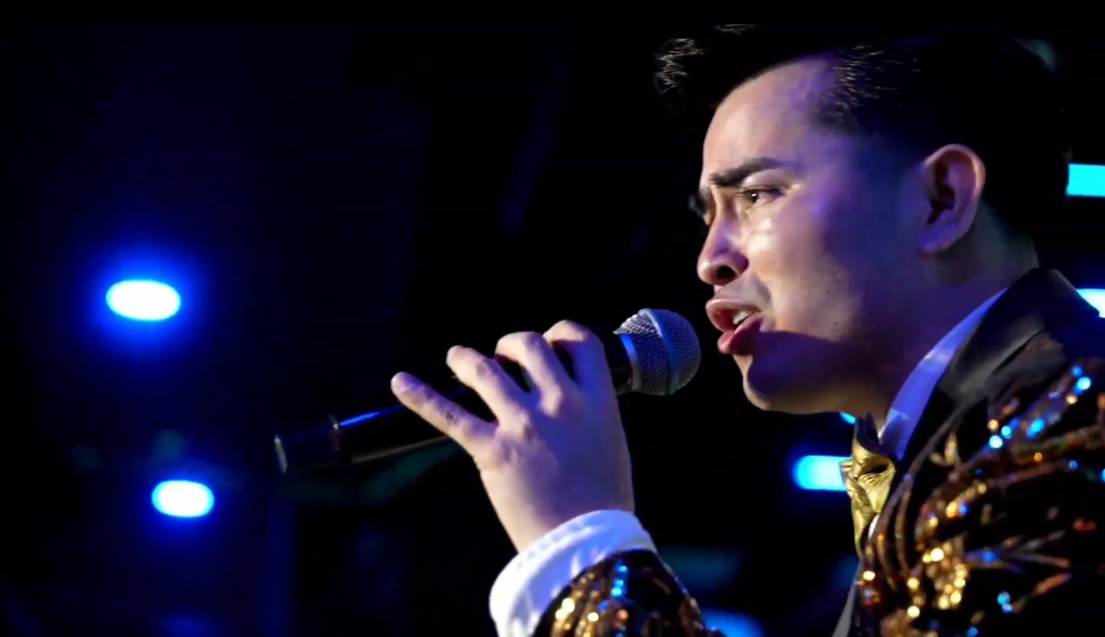 Man singing into a microphone on stage, wearing a shiny, glittery jacket with blue and gold accents, under blue stage lights. La voz Guacalito, Alberto Jimenez cantante latino