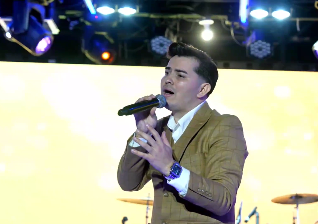 A male singer in a tan suit and white shirt singing into a microphone on stage with a bright yellow background and stage lights overhead.El Guacalito, La Voz el Guacalito