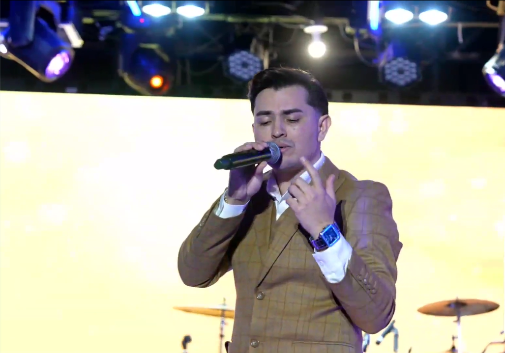 A man in a brown suit singing on stage with a microphone, with drum equipment visible in the background under stage lights.El Guacalito, La Voz el Guacalito