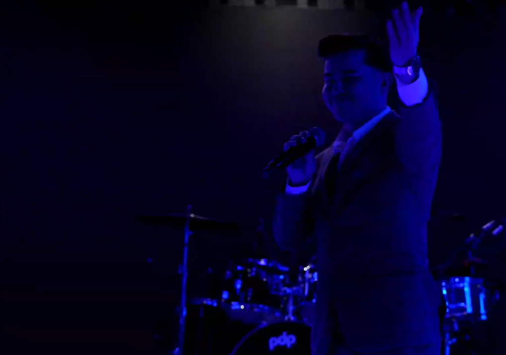 A man in a suit singing into a microphone on stage, silhouetted against blue stage lighting, with drums in the background.El Guacalito, La Voz el Guacalito