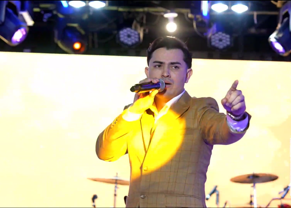 A male performer wearing a tan suit singing into a microphone on stage, with a drum set visible in the background and stage lights overhead.El Guacalito, La Voz el Guacalito
