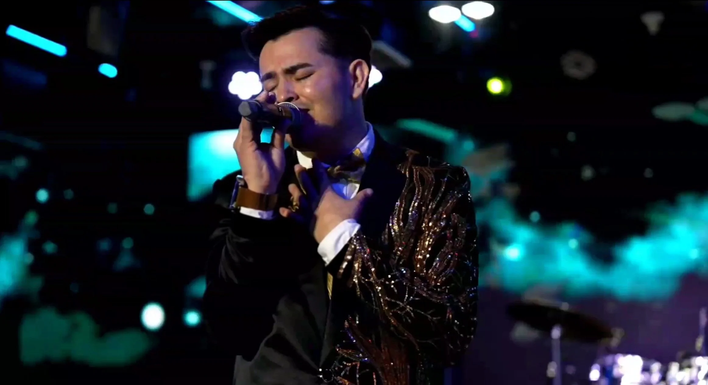A man in a black jacket with gold and black patterns, wearing a white shirt and bow tie, passionately singing into a microphone with his eyes closed, on stage with blue and purple lights. La voz Guacalito, Alberto Jimenez cantante latino