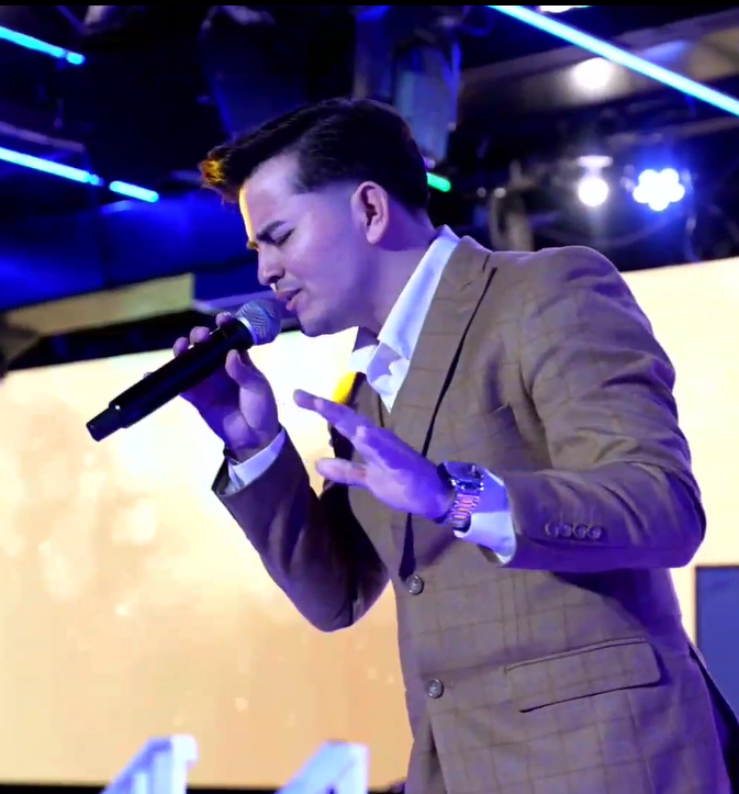 A man in a brown blazer singing into a microphone on stage with colorful lighting.
El Guacalito, La Voz el Guacalito