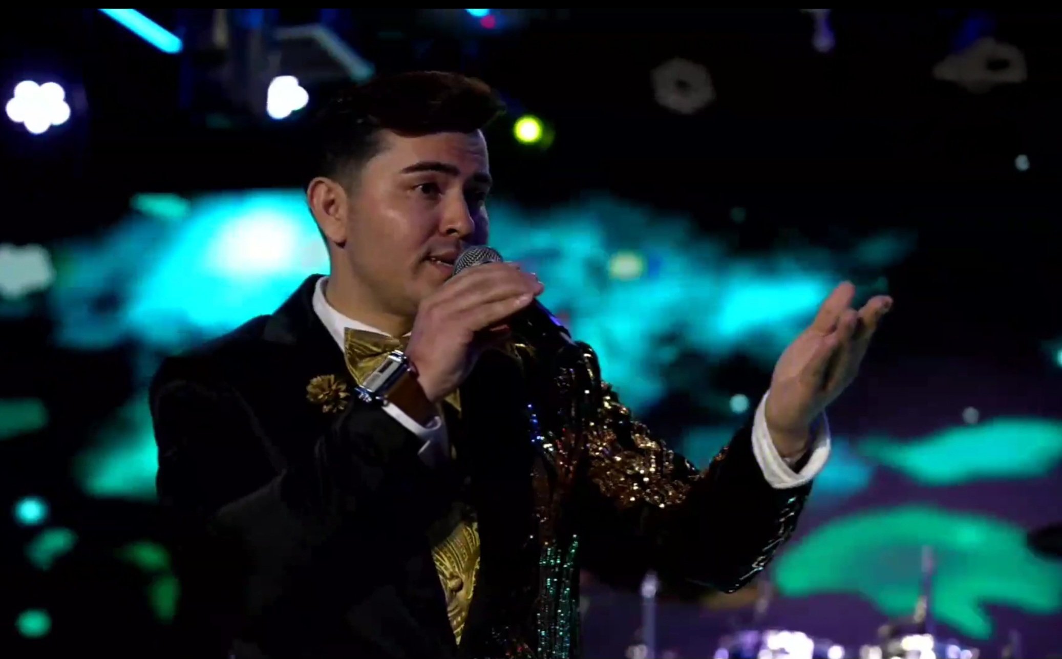 A man wearing a black jacket with gold embellishments and a gold bowtie is speaking into a microphone on stage, with a colorful, blurred background. La voz Guacalito, Alberto Jimenez cantante latino
