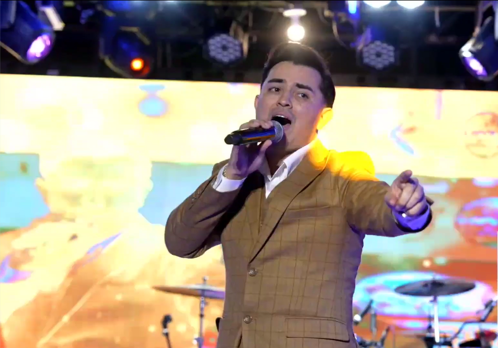 Male singer in a beige checkered suit singing into a microphone on stage with colorful lights and a drum set in the background El Guacalito, La Voz el Guacalito