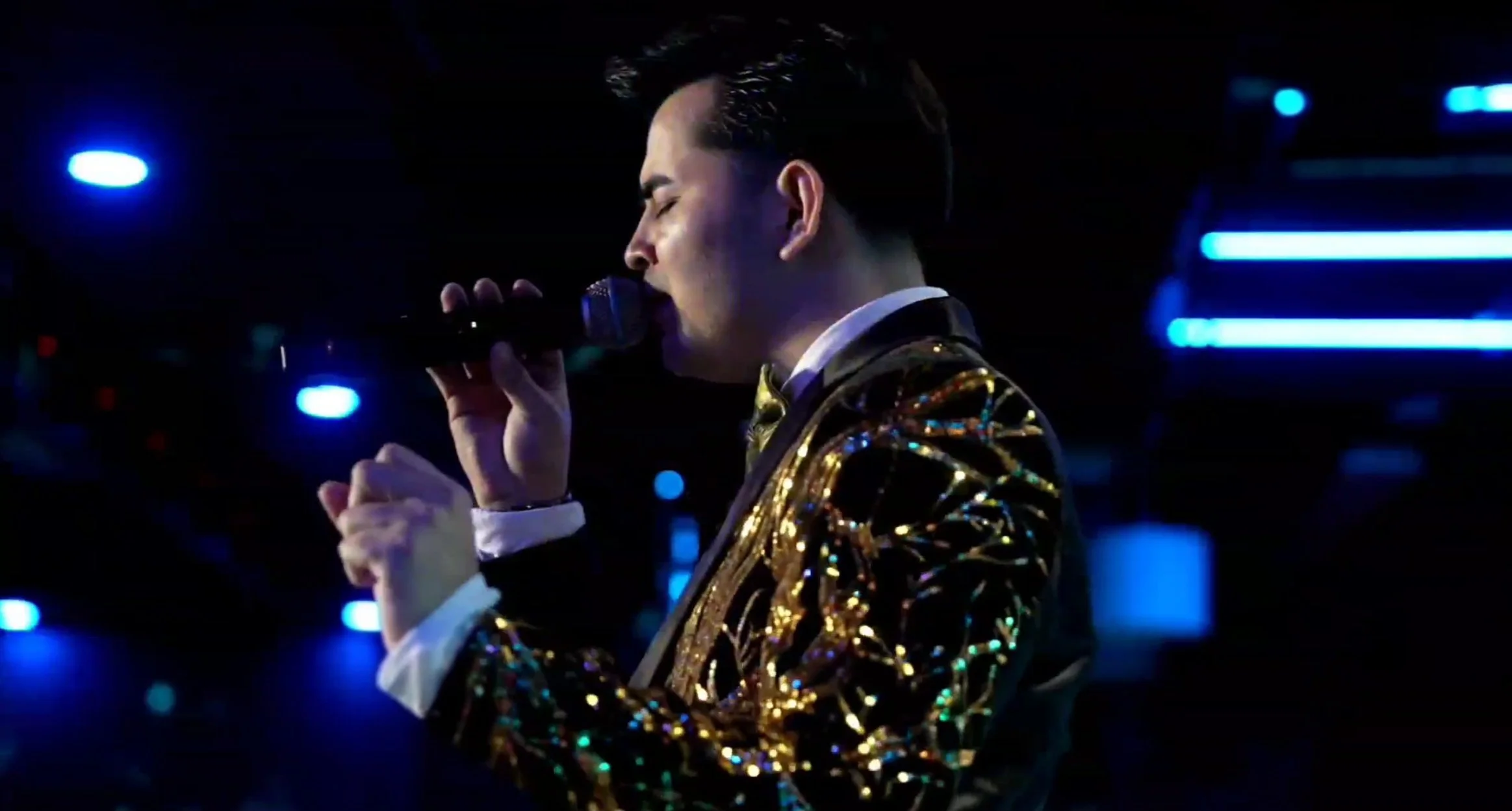 A male singer wearing a gold, sequined blazer performing with a microphone in a dimly lit venue with blue and purple lighting. La voz Guacalito, Alberto Jimenez cantante latino