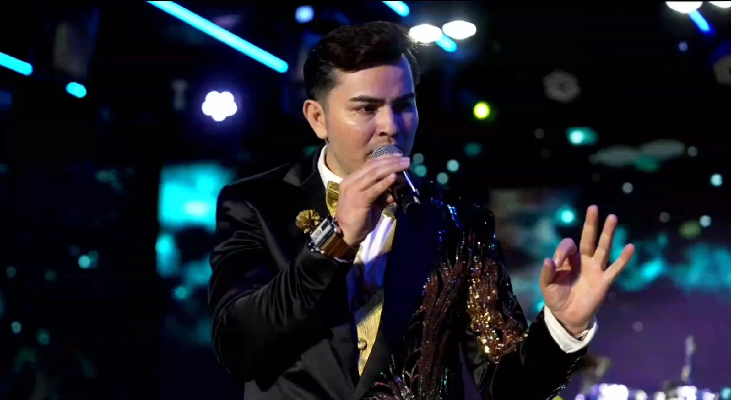 Man singing into a microphone with a hand gesture in a dark, colorful nightclub or concert venue. La voz Guacalito, Alberto Jimenez cantante latino