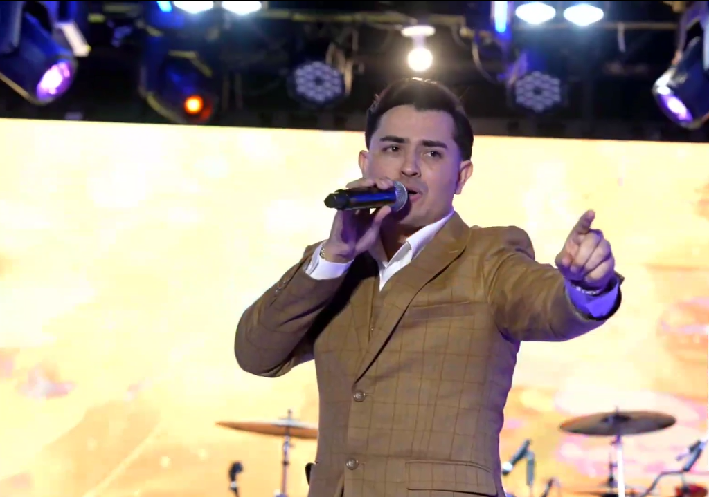 A man in a brown checkered suit holding a microphone and singing on stage, with colorful stage lights and drums in the background. El Guacalito, La Voz el Guacalito