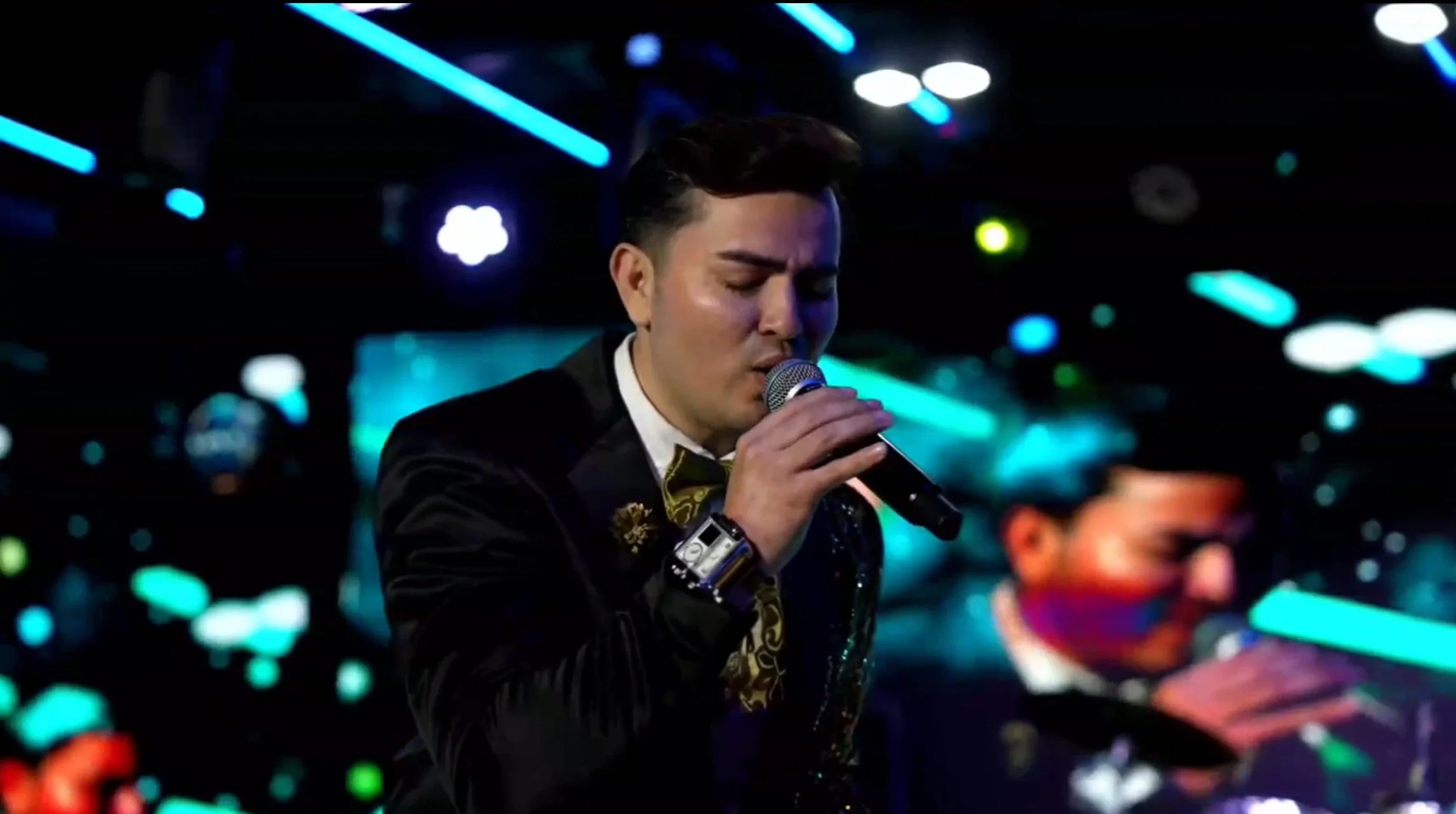 Man singing passionately with eyes closed, holding a microphone in a dimly lit venue with blue and green neon lights. La voz Guacalito, Alberto Jimenez cantante latino