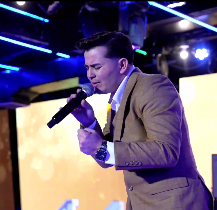 Man singing passionately into a microphone on stage, wearing a tan blazer and white shirt, with colorful stage lighting in the background.El Guacalito, La Voz el Guacalito