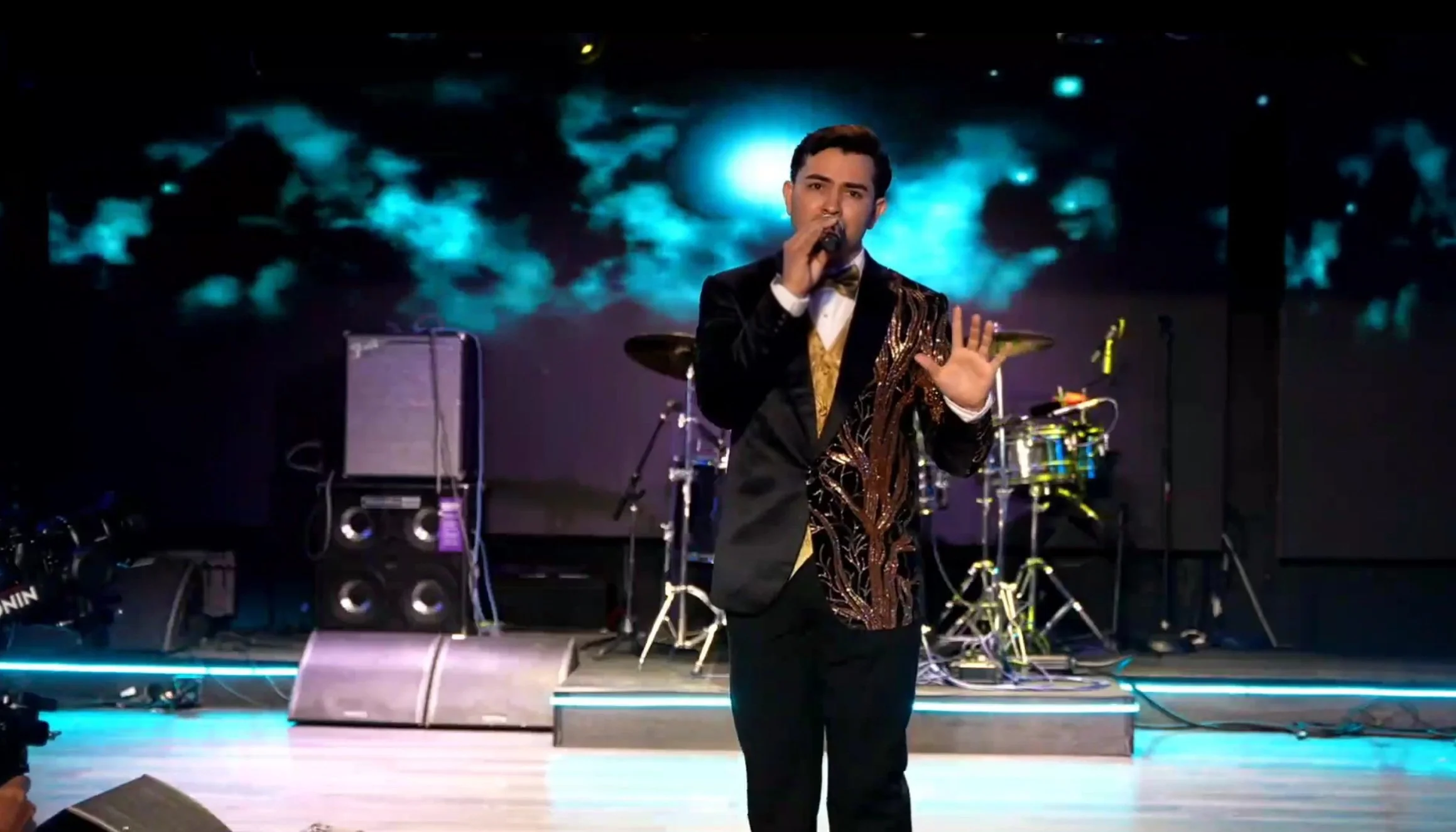 Man in a black suit with a shiny gold design, holding a microphone, performing on stage with musical instruments and blue lighting in the background. La voz Guacalito, Alberto Jimenez cantante latino