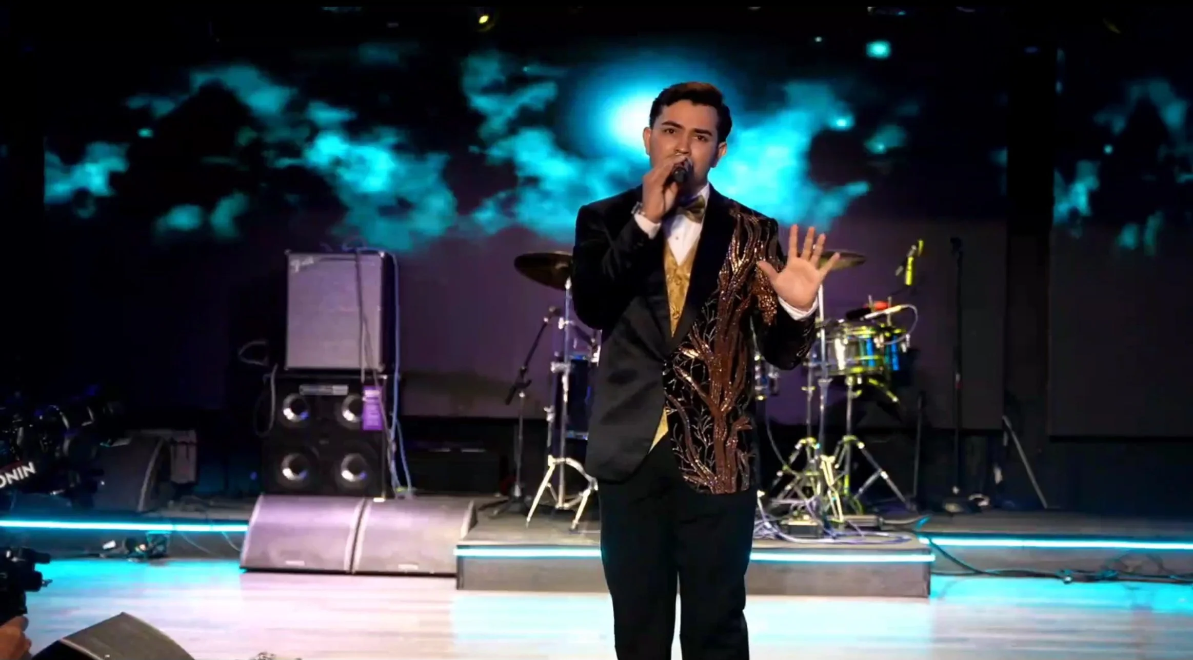 A man in a formal black suit with a gold and brown embroidered jacket performing on stage with a microphone, with musical instruments and a digital background behind him. La voz Guacalito, Alberto Jimenez cantante latino