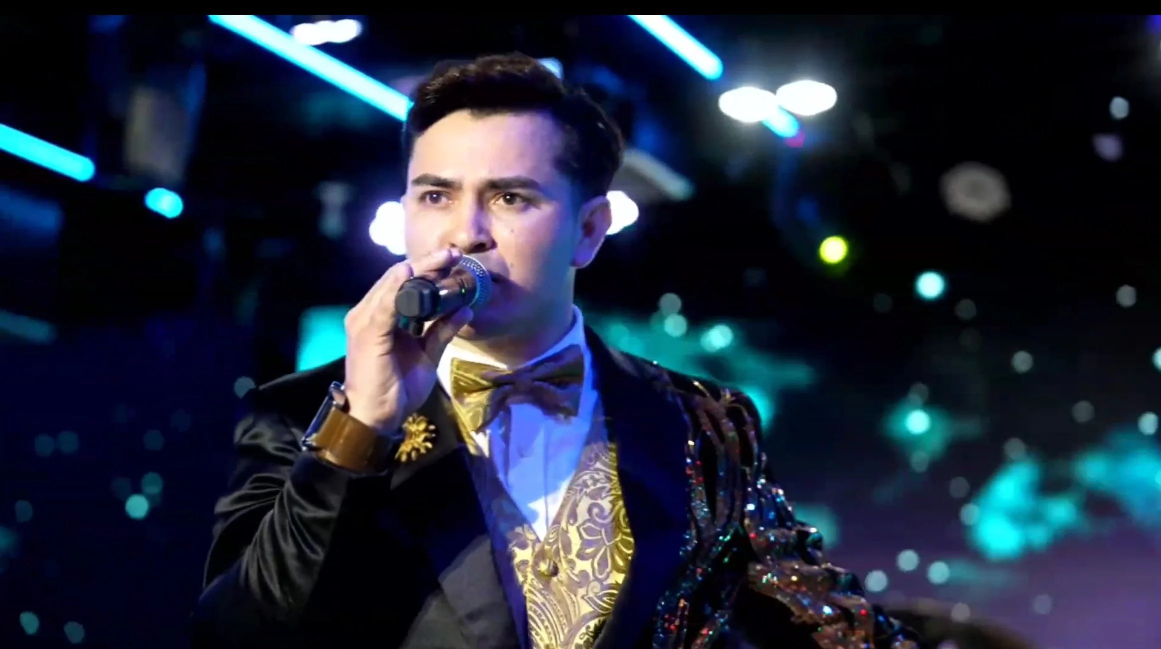 Young man performing on stage, holding a microphone, wearing a black jacket with gold accents and a bow tie, with colorful stage lights in the background. La voz Guacalito, Alberto Jimenez cantante latino
