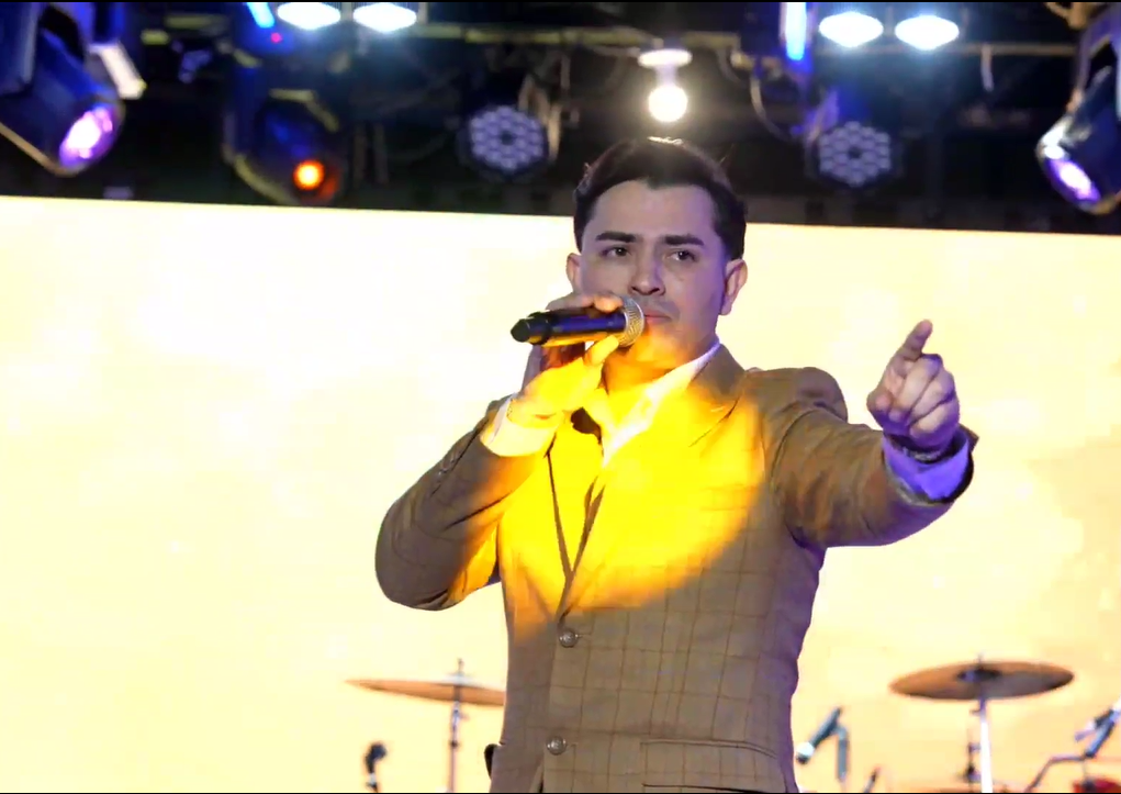 A male singer in a mustard yellow blazer performs on stage, holding a microphone, with a bright light illuminating him from the front and drums in the background. El Guacalito, La Voz el Guacalito