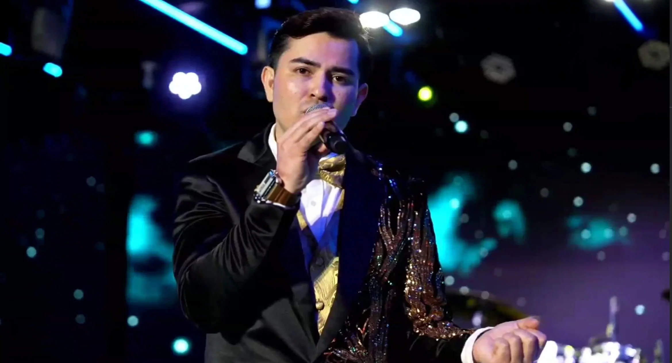 A male singer performing on stage with a microphone, wearing a black sequined blazer and formal attire, with colorful stage lights illuminating the background. La voz Guacalito, Alberto Jimenez cantante latino