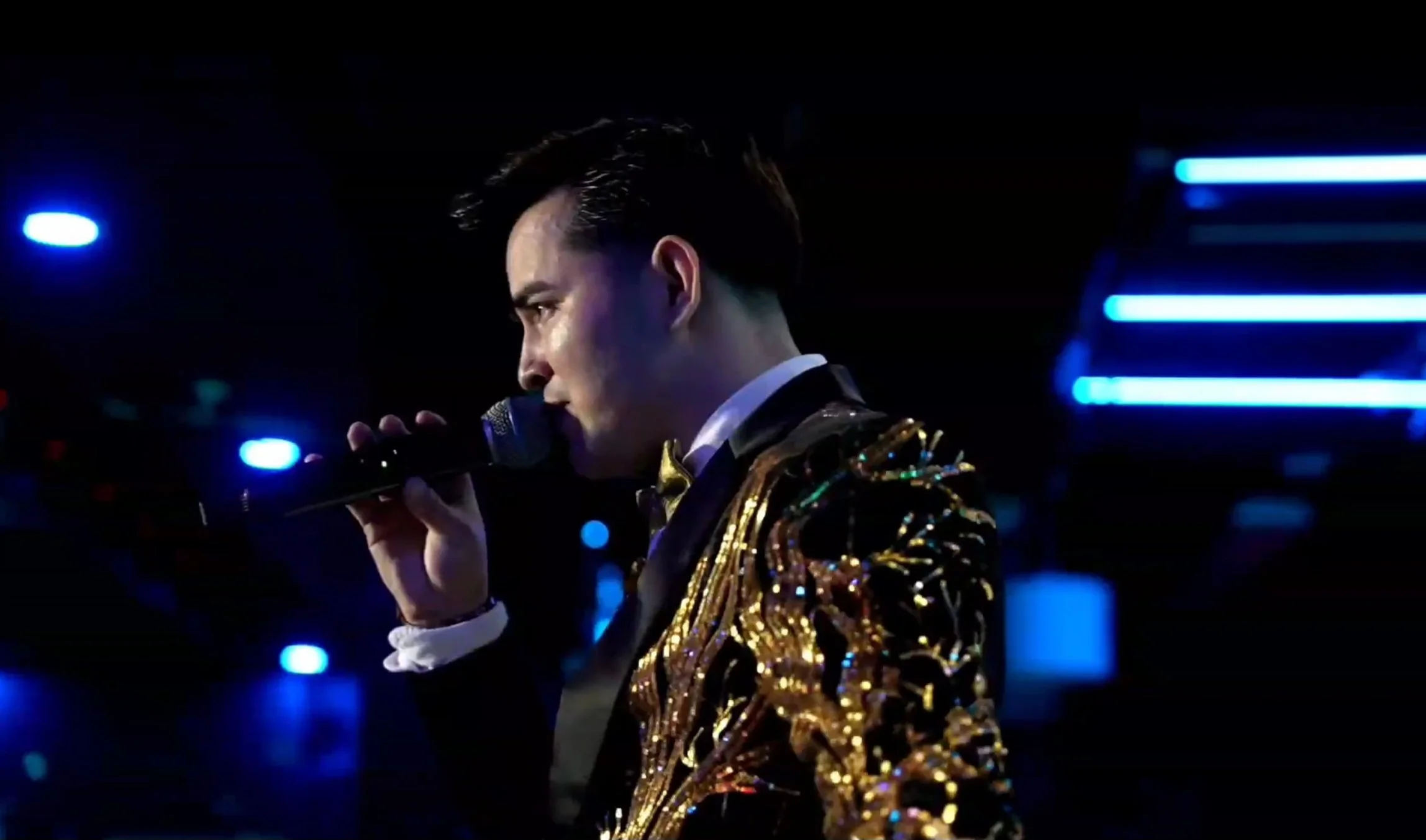 A man in a shiny, gold-sequined jacket holding a microphone and singing in a dark, neon-lit stage environment. La voz Guacalito, Alberto Jimenez cantante latino