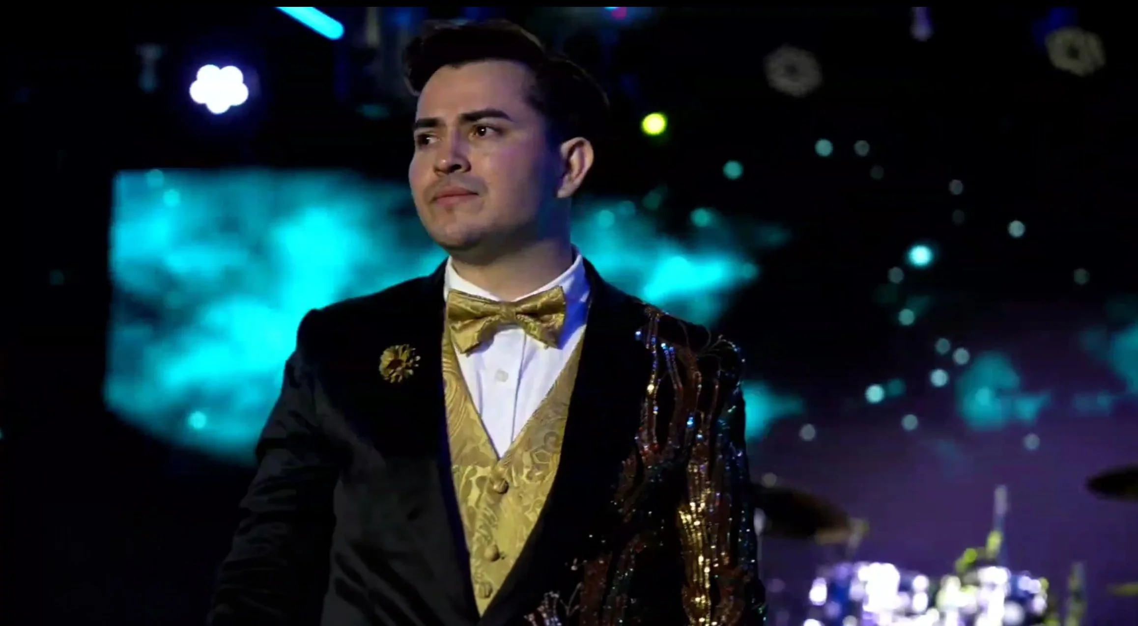 A man dressed in a black tuxedo with a gold vest and bow tie, standing on a stage with a colorful, starry background, during a performance or event. La voz Guacalito, Alberto Jimenez cantante latino
