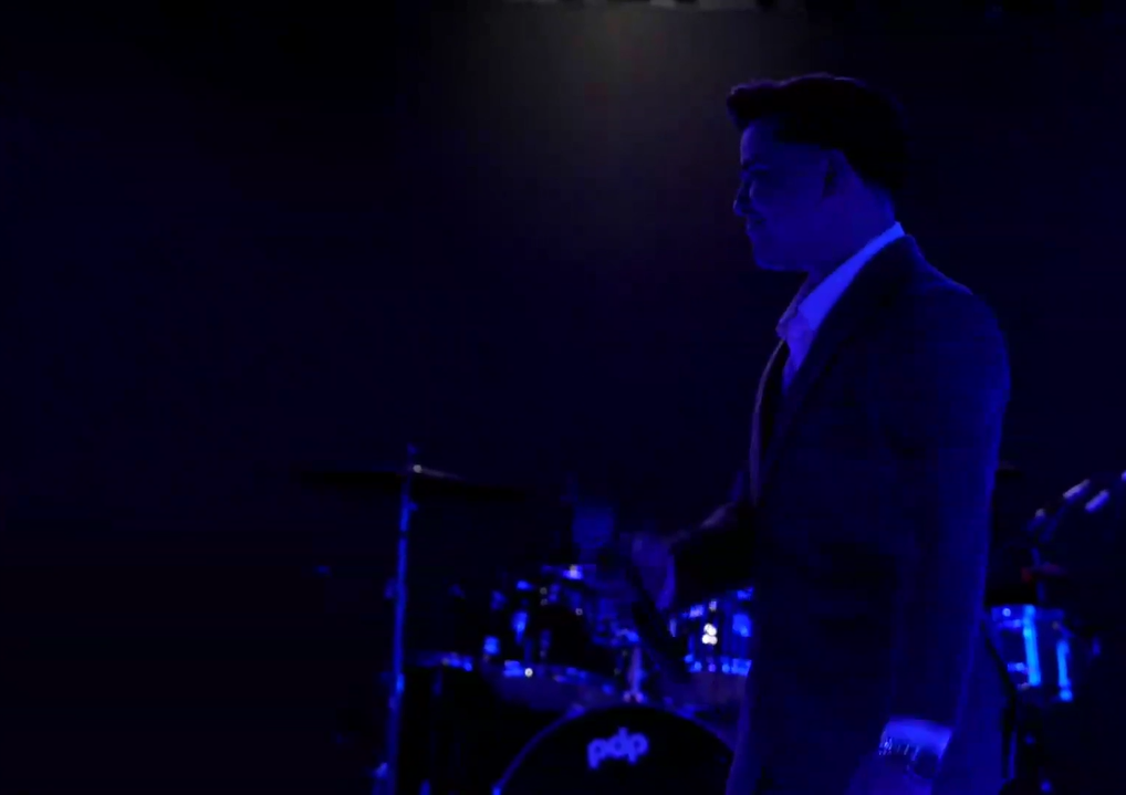 A man in a suit standing in a dark room with stage lighting, with a drum set in the background. El Guacalito, La Voz el Guacalito